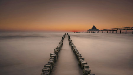 A tranquil image capturing a long wooden pier extending into a calm body of water during sunset. The...