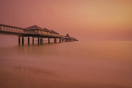 A tranquil scene of a pier extending into calm waters during a vibrant sunset. The sky is painted...