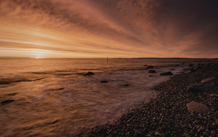 A serene sunset over a calm ocean with gentle waves lapping against the shore. A solitary sailboat...