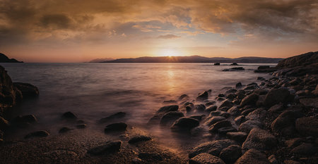 A serene sunset over a calm seascape with rocky foreground and distant islands.
