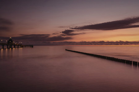 A serene image of a sunset over calm water with a pier extending into the distance. The sky is...