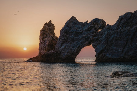 A breathtaking sunset casts warm hues over a coastal landscape featuring prominent rock arches. The sun dips below the horizon, creating a serene and picturesque scene.の写真素材
