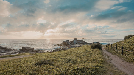 A serene coastal scene at sunset with cliffs overlooking the ocean, a dirt path, and lush greenery.の写真素材