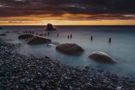 A serene sunset over a rocky beach with waves gently crashing against the shore. The sky is painted with hues of orange and purple, creating a dramatic backdrop.の写真素材