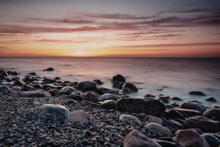 A serene sunset over a rocky beach with the sun dipping below the horizon, casting warm hues across the sky and reflecting off the calm sea.の写真素材