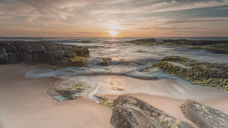 A serene coastal scene at sunset, featuring a rocky shoreline with waves gently lapping against the rocks. The sky is painted with hues of orange and pink as the sun dips below the horizon.の写真素材
