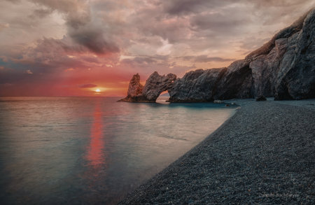 A serene sunset over a rocky coastline with calm waters reflecting the vibrant sky and clouds.の写真素材