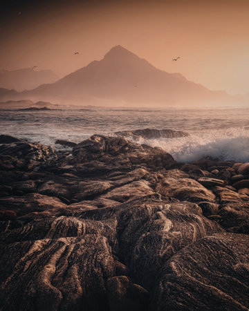 A serene coastal scene at sunset with rugged rocks in the foreground and a mountain in the background, with birds flying overhead.の写真素材