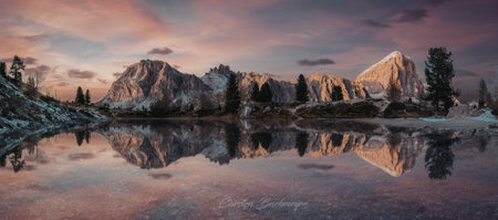 A serene landscape featuring snow-capped mountains at sunset, with their reflection perfectly mirrored in a calm lake. The sky is painted with hues of pink and orange, enhancing the tranquil atmosphere.の写真素材
