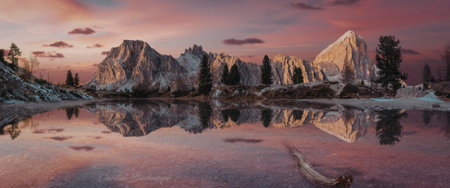 A breathtaking sunset casts a warm glow over snow-capped mountains, reflected perfectly in a calm lake surrounded by evergreen trees.の写真素材