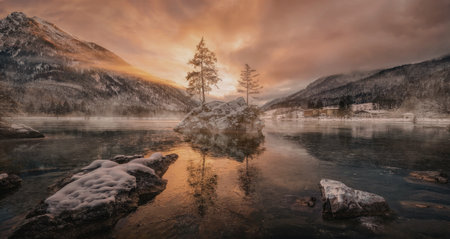 A serene winter landscape featuring a solitary tree on a small island in a calm lake, surrounded by snow-covered mountains and rocks, with a vibrant sunset casting warm hues across the sky.の写真素材