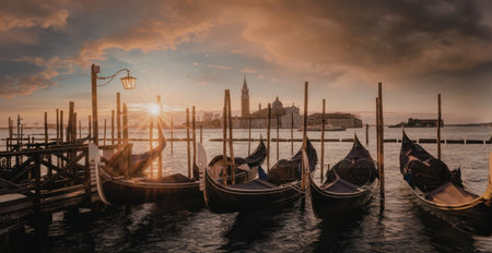 A serene sunset scene in Venice, featuring traditional gondolas moored at a wooden dock. The sky is painted with hues of orange and red, reflecting on the calm water, with the iconic cityscape in the background.の写真素材