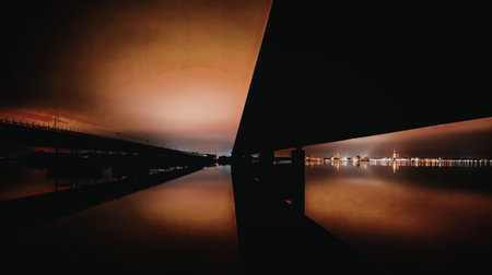 A serene image capturing the reflection of a vibrant sunset on a calm waterway, with a modern bridge...