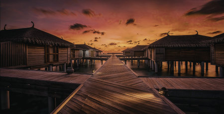A serene image of water villas situated over a calm body of water, with a vibrant sunset casting warm hues across the sky and reflections on the water.の写真素材