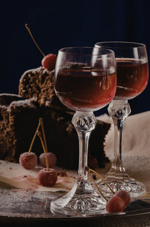 A close-up image of two elegant wine glasses filled with red wine, accompanied by a slice of chocolate cake and garnished cherries on a dark background.の写真素材