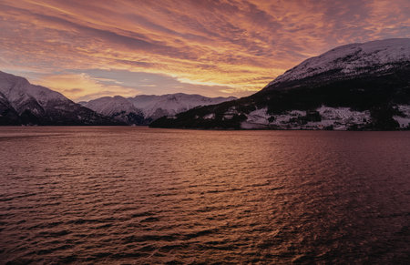 A serene landscape featuring a calm lake reflecting the vibrant hues of a sunset. Snow-covered mountains stand majestically in the background, creating a picturesque and tranquil scene.の写真素材