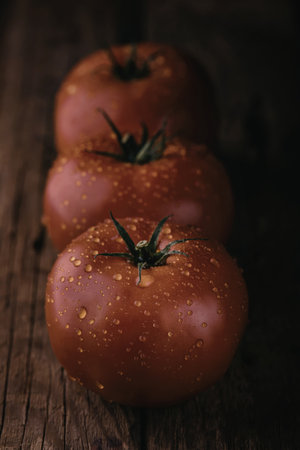 A close-up photograph of three ripe, red tomatoes arranged in a row on a rustic wooden surface. The tomatoes have visible water droplets on their skin and green stems attached.の写真素材