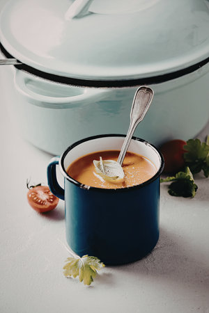 A close-up image of a blue mug filled with vibrant tomato soup, garnished with a basil leaf. A spoon rests in the mug, and a large pot is visible in the background. Fresh tomatoes and parsley are scattered around the mug on a light-colored surface.の写真素材