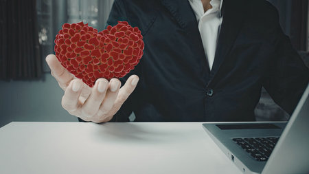A business professional in a suit is holding a red heart-shaped sculpture composed of interlocking chains. The individual is seated at a desk with a laptop in front of them, suggesting a blend of professionalism and emotional connection.の写真素材