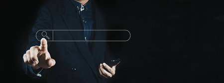 A close-up image of a businessman in a suit pointing at a virtual search bar with his finger,...