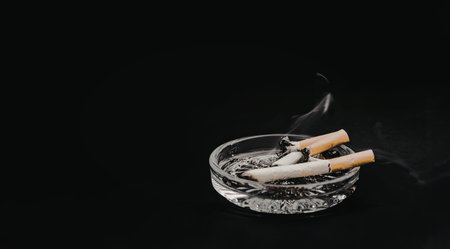 A clear glass ashtray containing a lit cigarette and cigarette butts, with smoke rising from the lit...