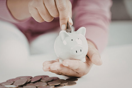 A child is carefully placing coins into a white piggy bank, demonstrating the concept of saving...