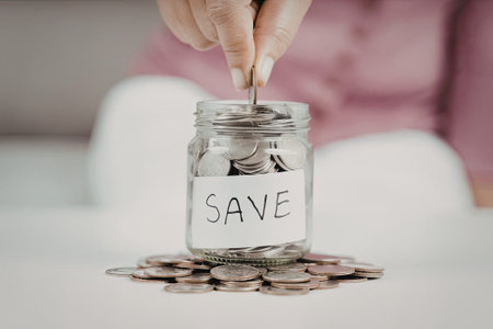 A person is adding a coin to a glass jar labeled 'SAVE', which is filled with coins. Additional...