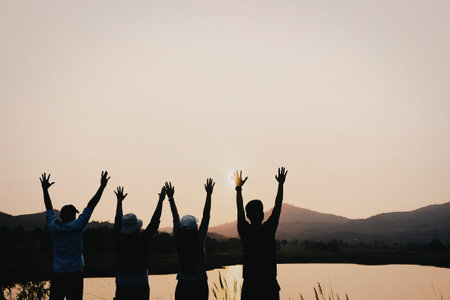 A group of people standing by a lake, raising their hands in celebration as the sun sets in the background, creating a serene and joyful atmosphere.の写真素材