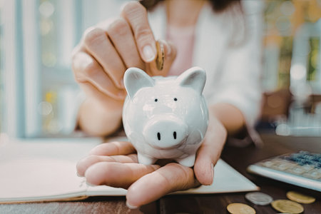 A person's hand is holding a white piggy bank while another hand is placing coins into it. The scene...
