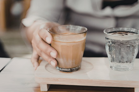 A person holding a glass cup filled with coffee, accompanied by another glass of water on a wooden tray.の写真素材
