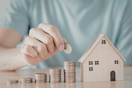 An image depicting a hand placing a coin on top of a stack of coins next to a wooden house model....