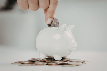 A close-up image of a hand placing a coin into a white piggy bank, surrounded by scattered coins.の写真素材