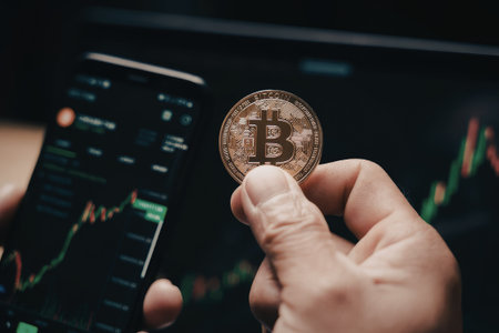 An image of a hand holding a physical Bitcoin coin in front of a smartphone displaying a financial chart with cryptocurrency data.の写真素材