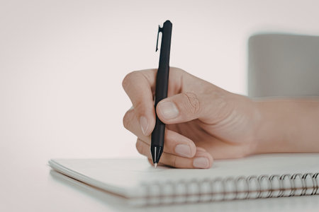 A close-up image of a hand holding a black pen, writing in a spiral-bound notebook. The background...