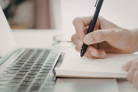 A close-up image of a person's hand writing in a spiral-bound notebook with a pen, while a laptop is...