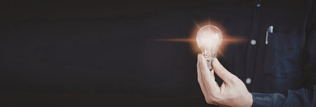 A close-up image of a hand holding a glowing light bulb against a dark background. The light bulb...