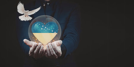 A pair of hands gently holding a glowing heart with blue and yellow colors, symbolizing peace and unity, with a white dove flying nearby.の写真素材