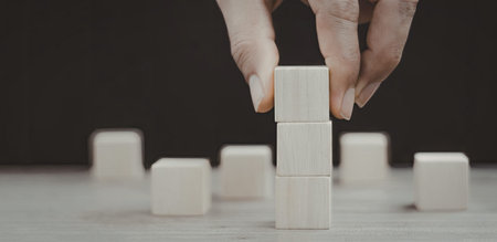 A close-up image of hands carefully stacking wooden blocks on a flat surface, with additional blocks scattered around.の写真素材
