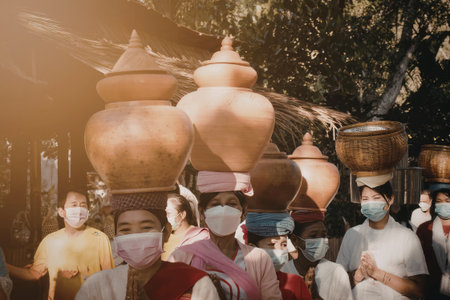 A group of people wearing masks and traditional attire are seen carrying large ceramic pots on their heads as part of a cultural or religious procession. The scene is set outdoors with sunlight filtering through trees in the background.の写真素材