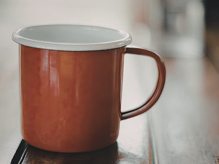 A close-up photograph of a red enamel mug with a white rim and handle, placed on a wooden surface....