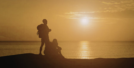 A romantic couple stands on the beach, their silhouettes framed against a vibrant sunset over the ocean. The sun casts a golden glow on the water, creating a serene and picturesque scene.の写真素材