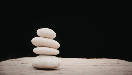 A stack of four white stones balanced on a wooden surface against a dark background. The image...