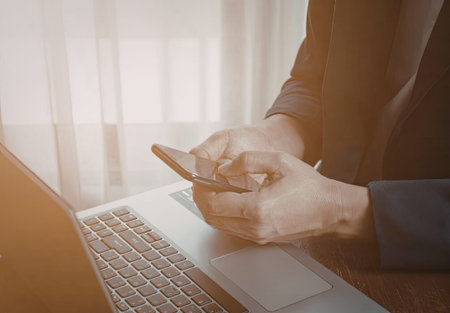 A person is holding a smartphone in one hand while using a laptop on a table. The scene is well-lit...