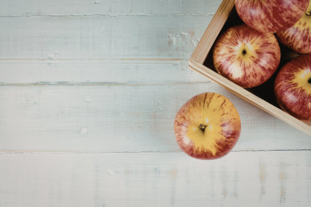 A wooden box filled with fresh, red apples sits on a white wooden surface. One apple has rolled out of the box, resting nearby.の写真素材
