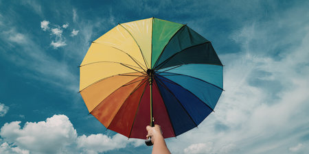 A hand holding a vibrant, multicolored umbrella stands out against a bright blue sky with scattered clouds.の写真素材