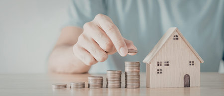 A close-up image of a hand placing a coin on top of a stack of coins next to a small wooden model...