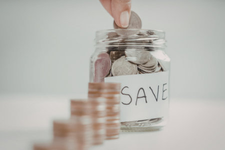 A hand is shown placing a coin into a clear glass jar labeled 'SAVE'. The jar is filled with various coins, and additional coins are stacked in front of it.の写真素材