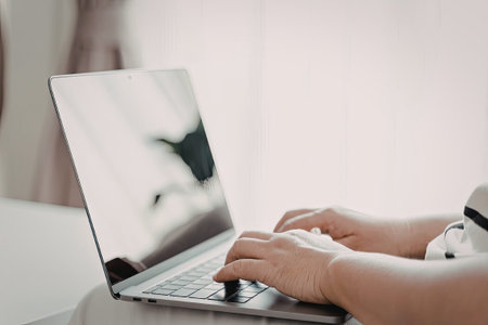 A close-up image of a person's hands typing on a laptop keyboard. The laptop is placed on a table,...