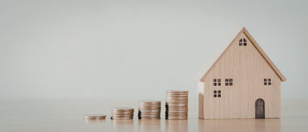 A small wooden model of a house is placed next to stacks of coins, symbolizing real estate...