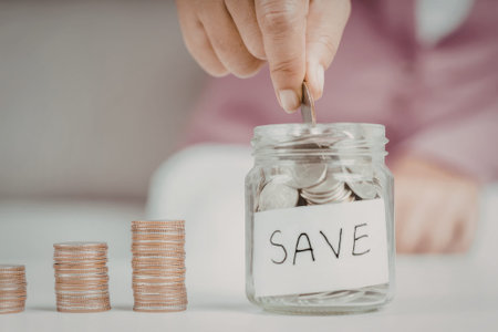 A person is depicted placing a coin into a glass jar labeled 'SAVE'. The jar is surrounded by stacks of coins, emphasizing the act of saving money.の写真素材